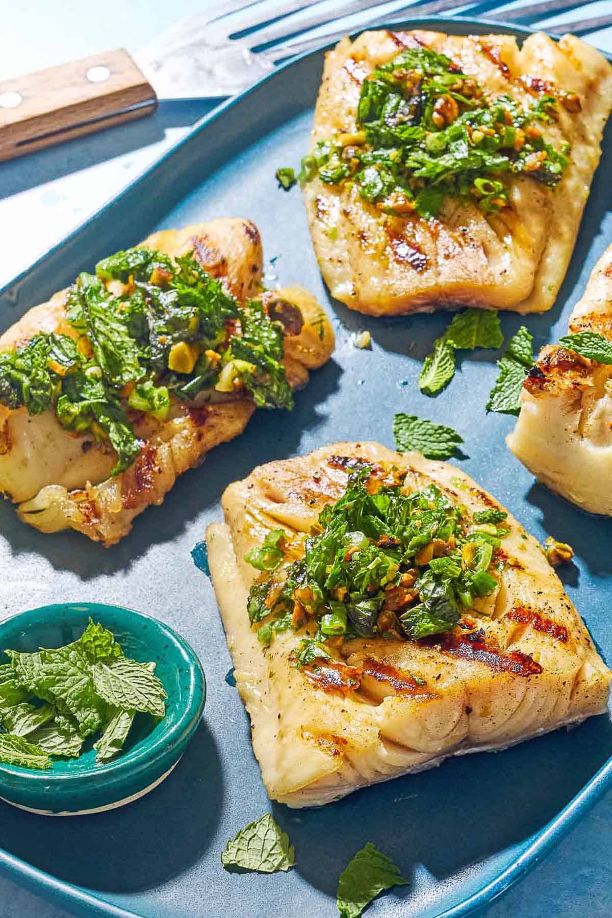 A close up of 3 grilled cod filets topped with pistachio-herb salsa on a serving platter along with a small bowl of mint leaves. Next to this is a fish spatula.