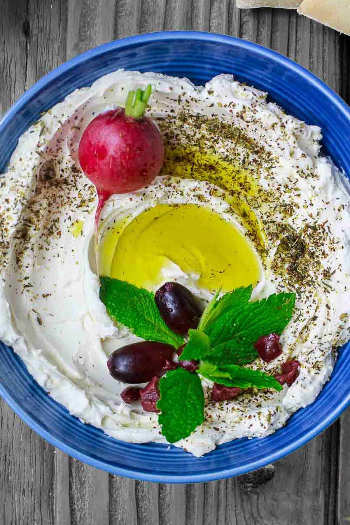 An overhead photo of homemade labneh in a bowl, garnished with herbs, olives, a radish and a drizzle of olive oil.