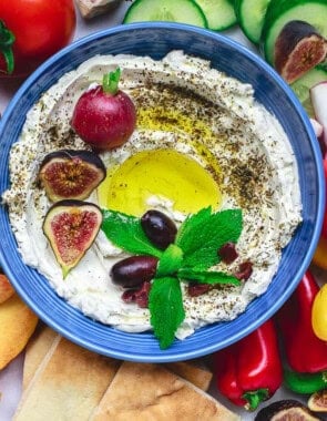 An overhead photo of homemade labneh in a bowl, garnished with herbs, olives, figs, a radish and a drizzle of olive oil surrounded by various fruits and raw vegetables and a bowl of za'atar.