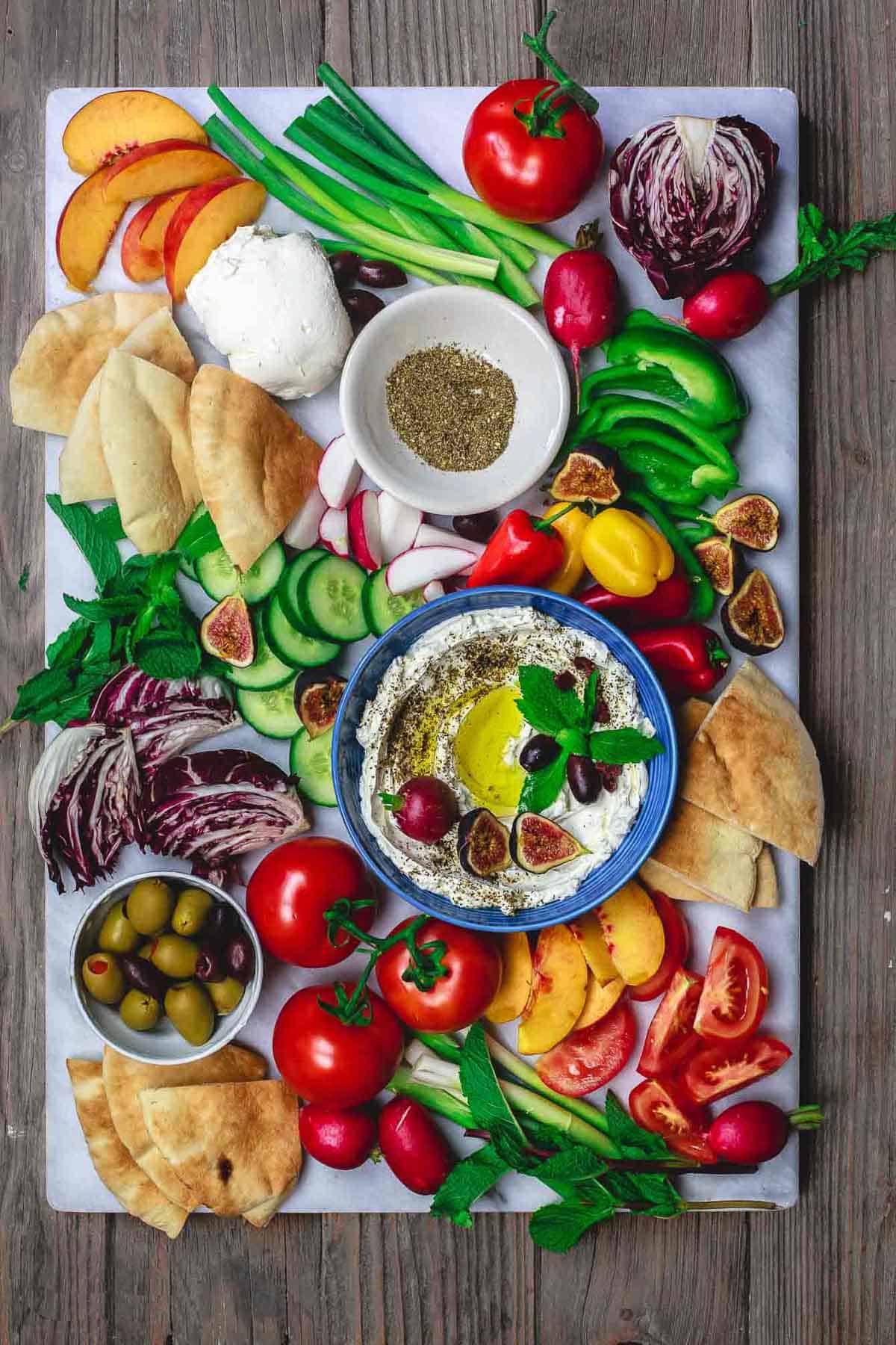 An overhead photo of homemade labneh recipe in a bowl, garnished with herbs, olives, figs, a radish and a drizzle of olive oil surrounded by, more labneh, various fruits and raw vegetables, pita bread wedges, a bowl of za'atar and a bowl of olives.