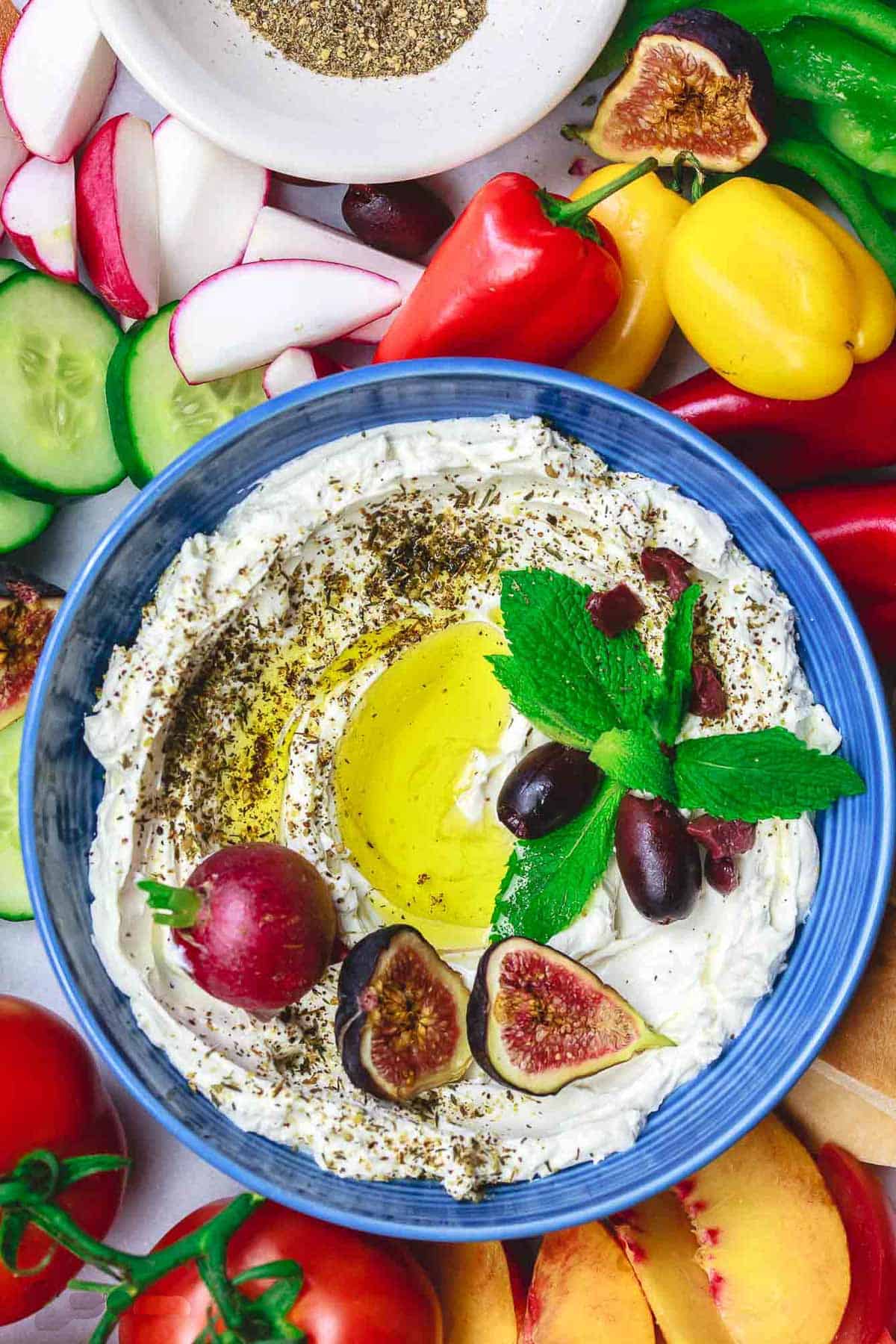 An overhead photo of homemade labneh in a bowl, garnished with herbs, olives, figs, a radish and a drizzle of olive oil surrounded by various fruits and raw vegetables.