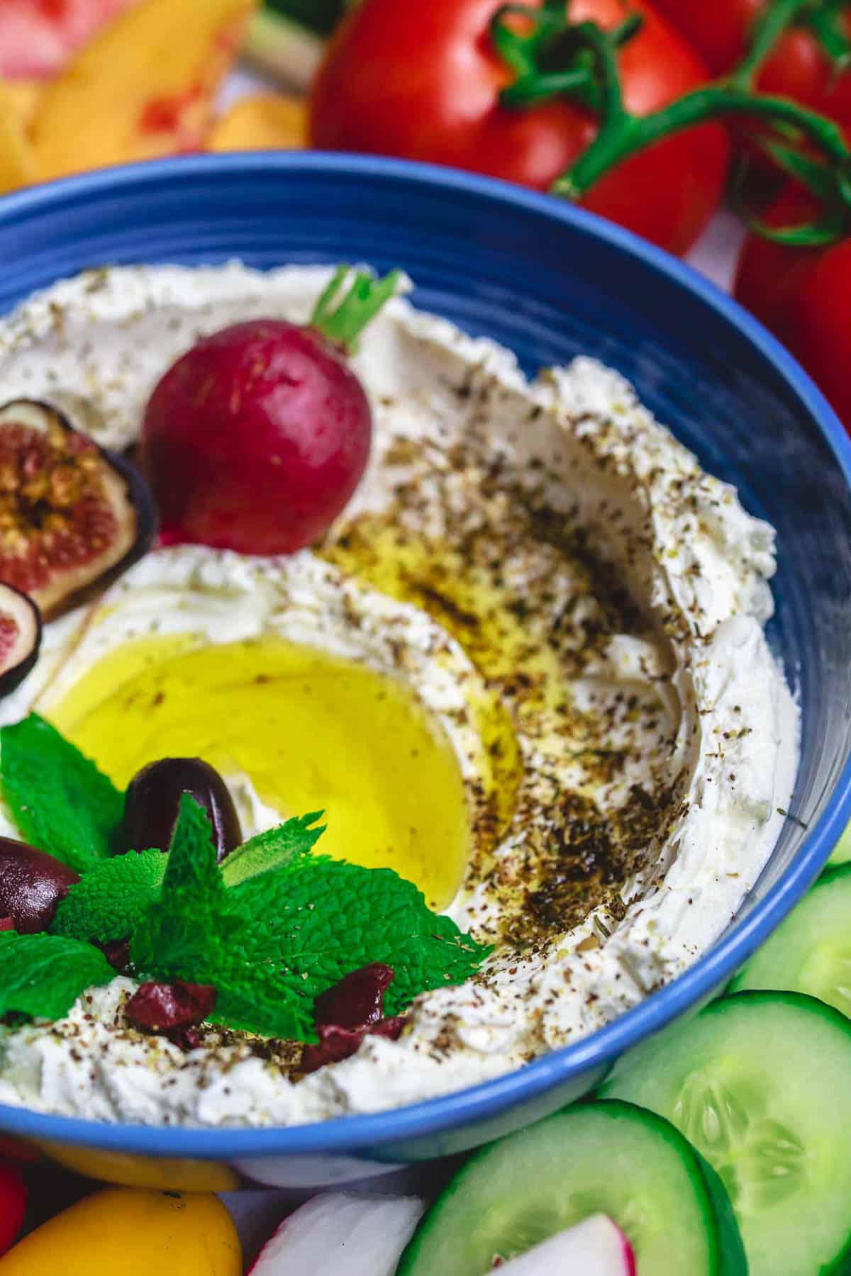 A close up of homemade labneh recipe in a bowl, garnished with herbs, olives, figs, a radish and a drizzle of olive oil surrounded by various raw vegetables.