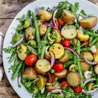 an overhead photo of Italian potato salad on a serving platter next to a stack of blue plates with forks, and a small bowl of chopped parsley.