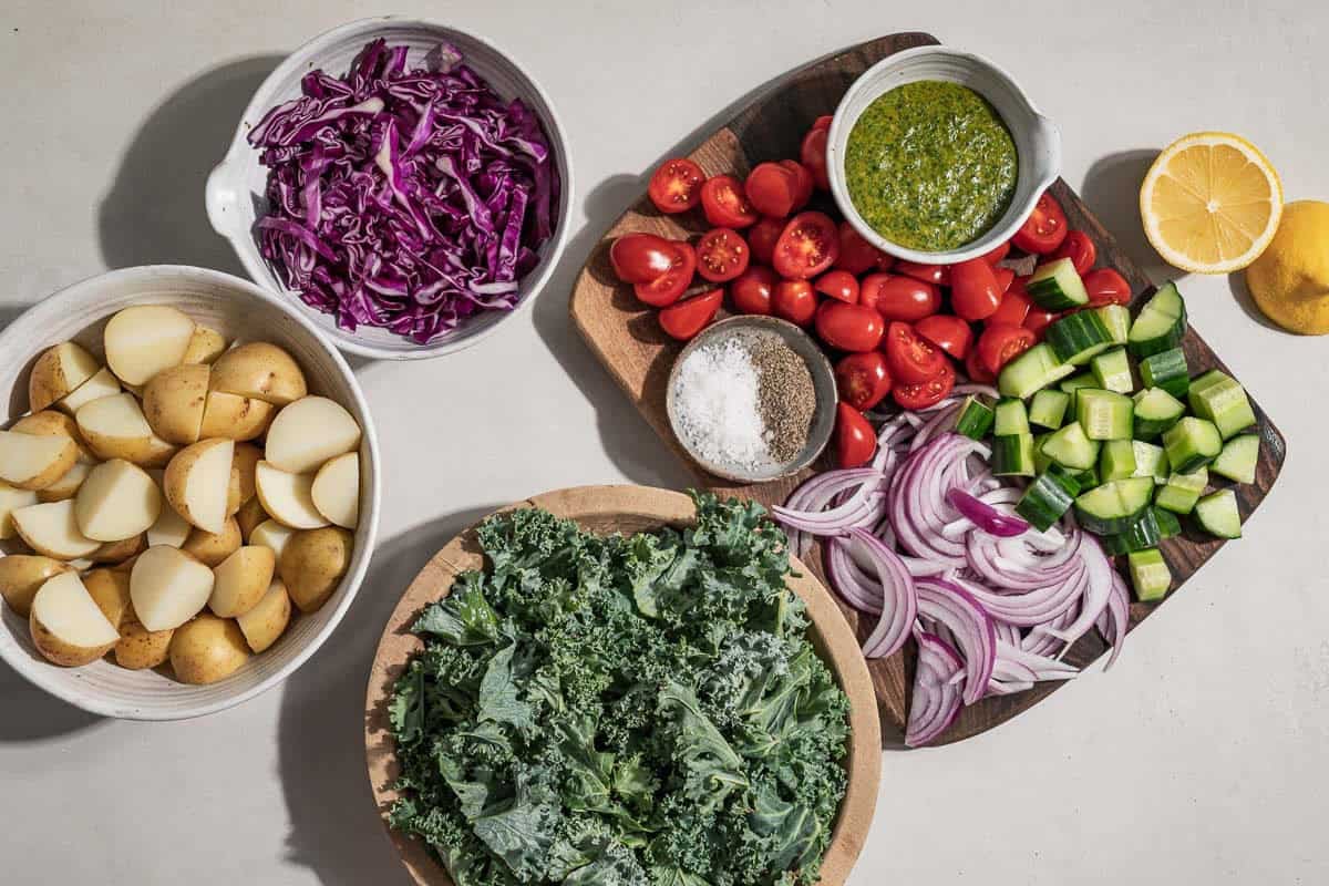 Ingredients for pesto potato salad including pesto, new potatoes, kale, cherry tomatoes, red onion, english cucumber lemon salt and black pepper.