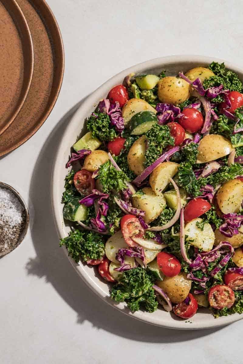 An overhead photo of a healthy potato salad in a serving bowl dressed in a lemon basil pesto dressing. Next to this is a stack of 2 plates and a small bowl of salt and black pepper.