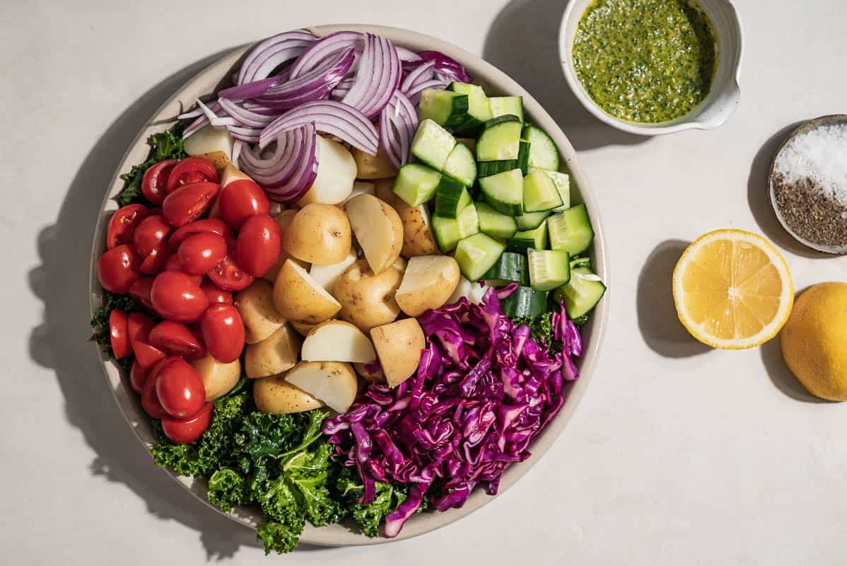 An overhead photo of the ingredients for a pesto potato salad in a serving bowl just before being mixed together. Next to this is a bowl of pesto, 2 lemon halves, and a small bowl of salt and pepper.