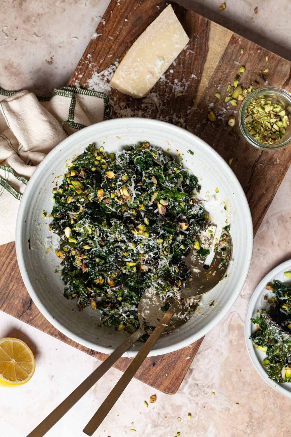 An overhead photo of kale salad in a large bowl with serving utensils on a cutting board. Next to this is a jar of chopped pistachios, a lemon half, a partial block of parmesan cheese, a cloth napkin, and a plate containing a serving of salad.