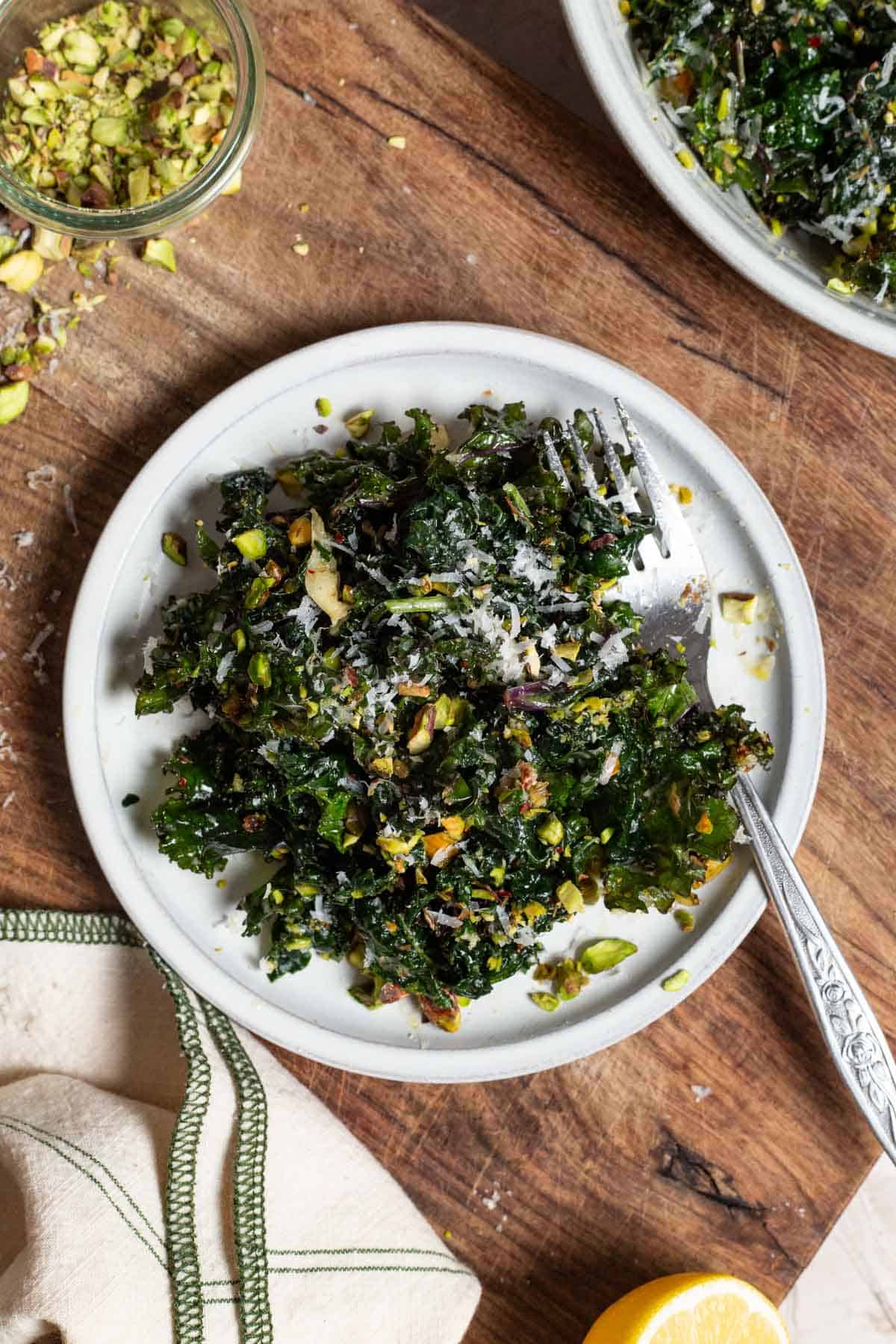 An overhead photo of a serving of kale salad on a plate with a fork on a cutting board. Next to this is a jar of chopped pistachios, a lemon half, a cloth napkin, and the rest of the salad in a large bowl.