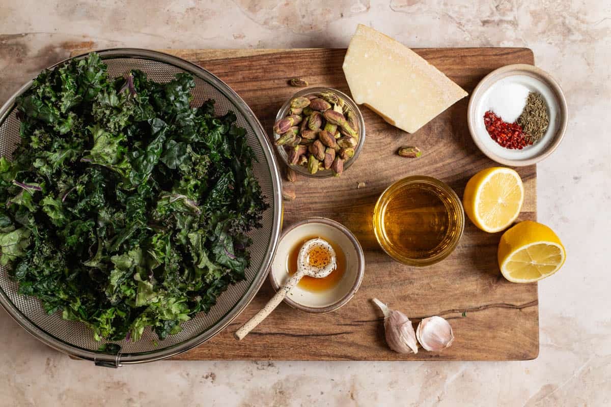 ingredients for kale salad including kale in a colander, lemon, olive oil, garlic, thyme, salt, aleppo pepper, pistachios, parmesan cheese and honey.