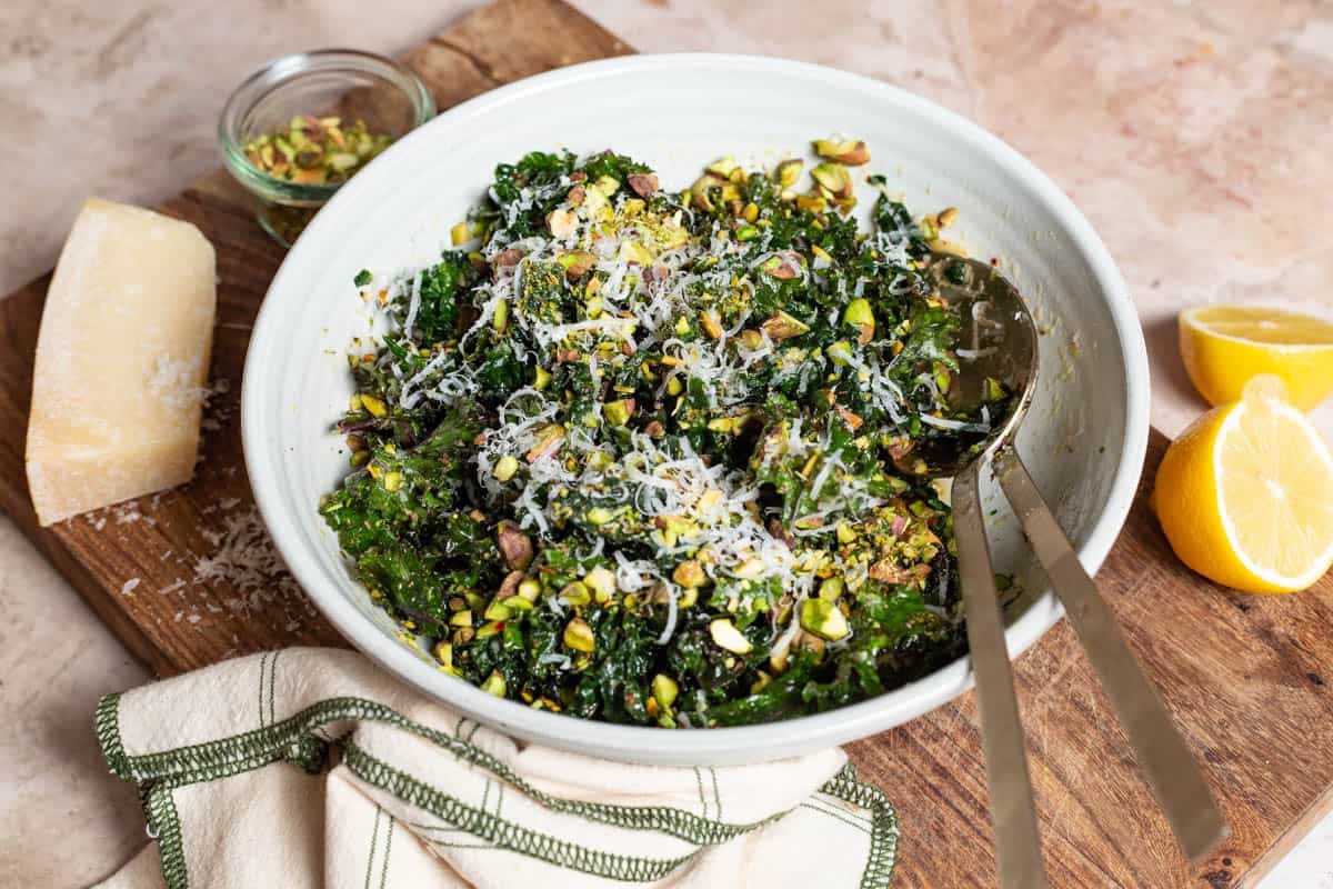 Kale salad in a large bowl with serving utensils on a cutting board. Next to this is a partial block of parmesan cheese, 2 lemon halves, a jar of chopped pistachios and a cloth napkin.