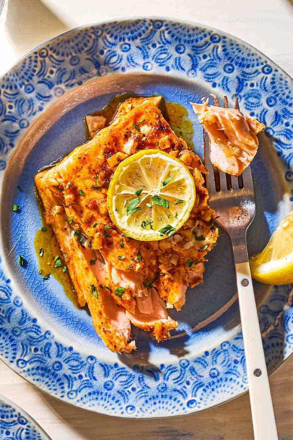 An overhead close up photo of a serving of the lemon garlic salmon topped with a lemon wheel and parsley on a plate with a fork.