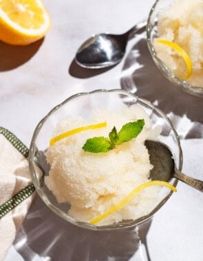 Two bowls of lemon sorbet, one with a spoon, and one next to a spoon. Next to these are 2 lemon wheels and a kitchen towel.