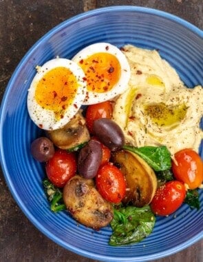 A Mediterranean breakfast bowl with a soft-boiled egg, hummus and sauteed vegetables on a blue plate.