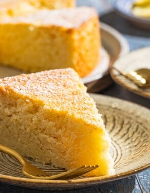 A close up of a slice of lemon ricotta cake on a plate with a fork with various other table settings in the background.