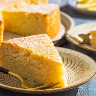 A close up of a slice of lemon ricotta cake on a plate with a fork with various other table settings in the background.