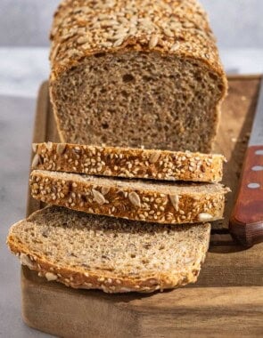 A close up photo of a partially sliced whole wheat bread loaf behind 3 slices of the bread on a cutting board with a knife.