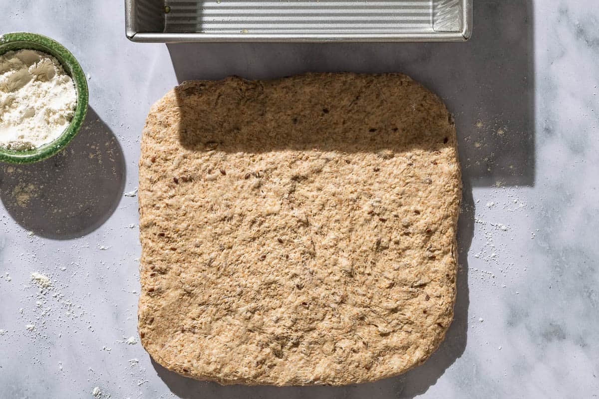 An overhead photo of whole wheat bread dough shaped into a large square next to a small bowl of flour and a loaf pan.