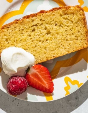 A close up of a slice of yogurt cake on a plate with a dollop of greek yogurt, a strawberry and a raspberry.