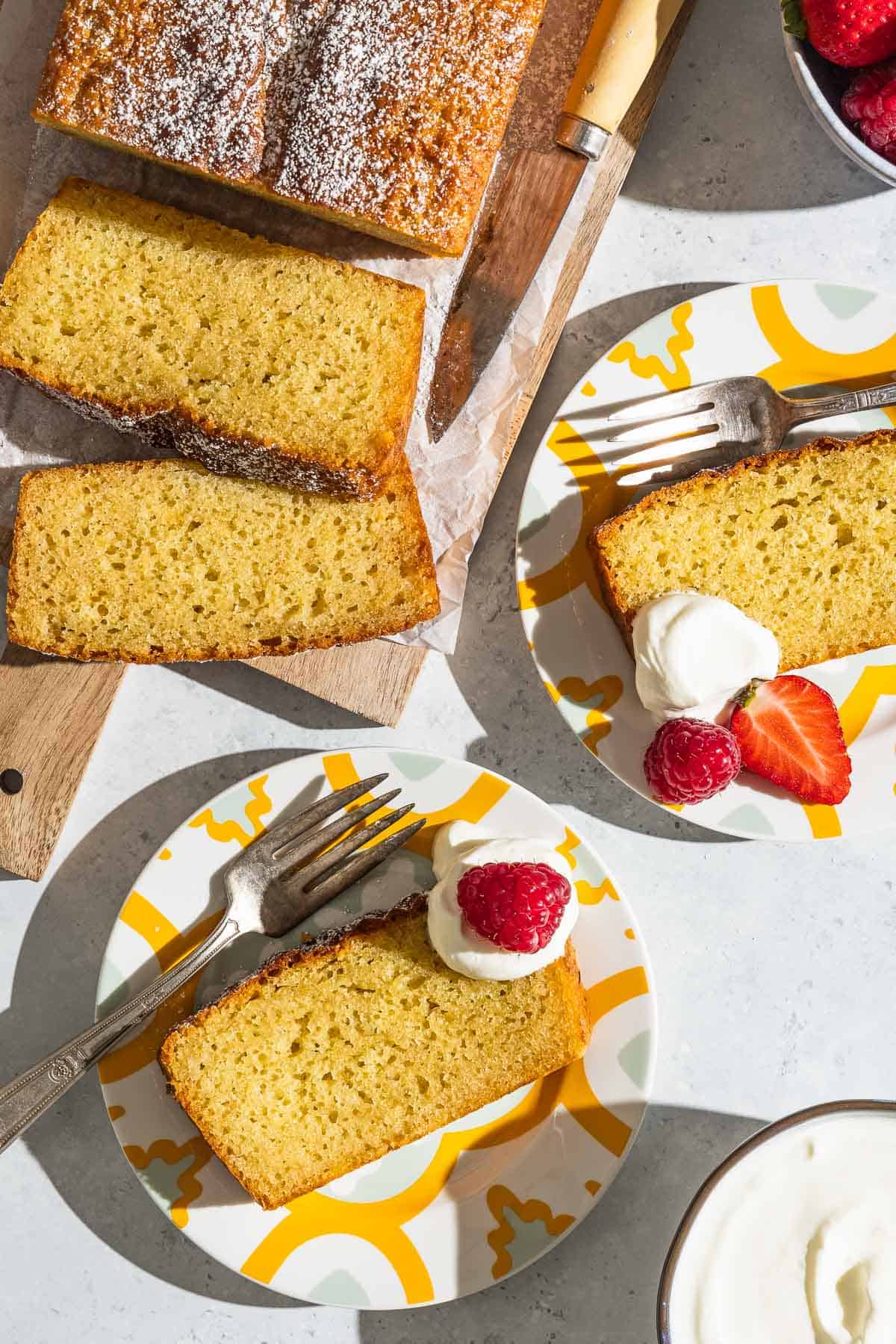 An overhead photo of yogurt cake cut into slices on a parchment lined cutting board with a knife, two slices of yogurt cake on 2 plates, each with a dollop of greek yogurt, some berries and a fork, and a bowl of greek yogurt.