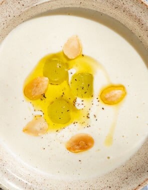 An overhead photo of ajo blanco white gazpacho garnished with green grapes, toasted almonds, olive oil and black pepper with a spoon next to it.