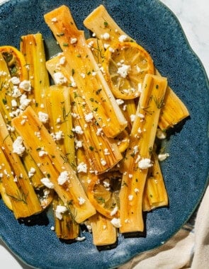 An overhead photo of braised leeks garnished with dill, crumbled feta and lemon slices on a blue serving platter.