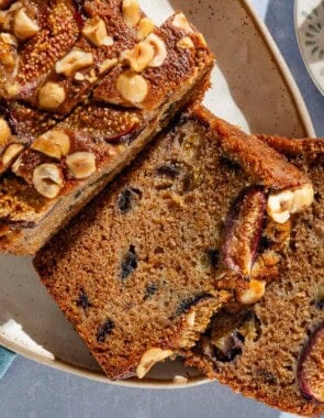 An overhead photo of a loaf of partially sliced whole wheat fig bread on a serving plate. Next to this are bowls of hazelnuts and figs.