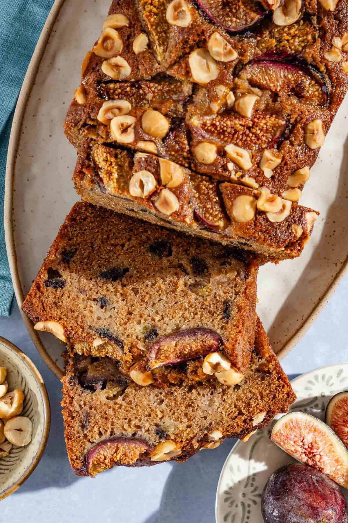 A loaf of partially sliced whole wheat fig bread on a serving plate. Next to this are bowls of hazelnuts and figs.