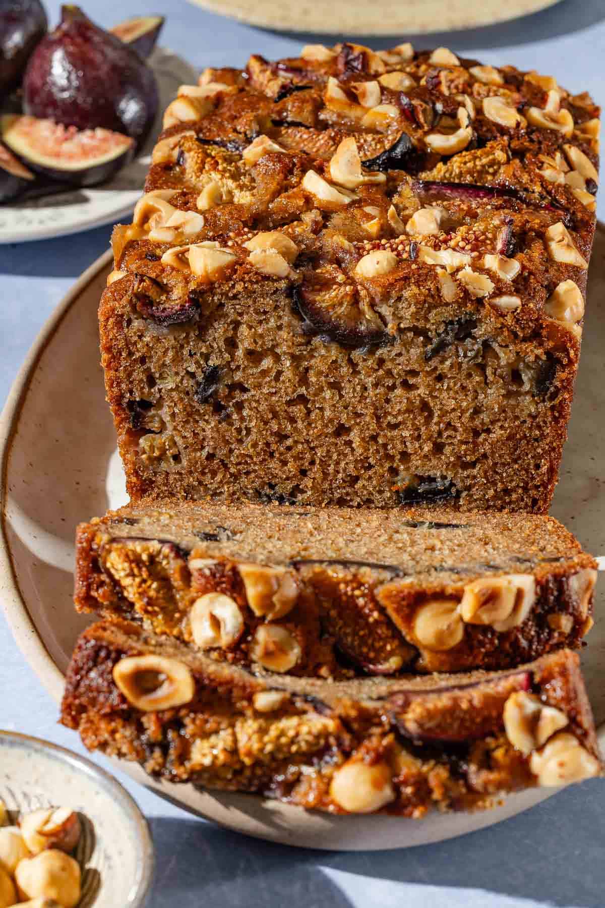 A close up of a loaf of partially sliced whole wheat fig bread on a serving plate. Next to this are bowls of hazelnuts and figs.