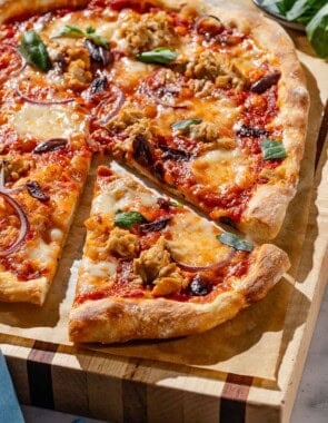 A close up photo of tuna pizza on a piece of parchment paper on a wooden cutting board, with one slice cut. Next to this is a cloth napkin and a bowl of basil.