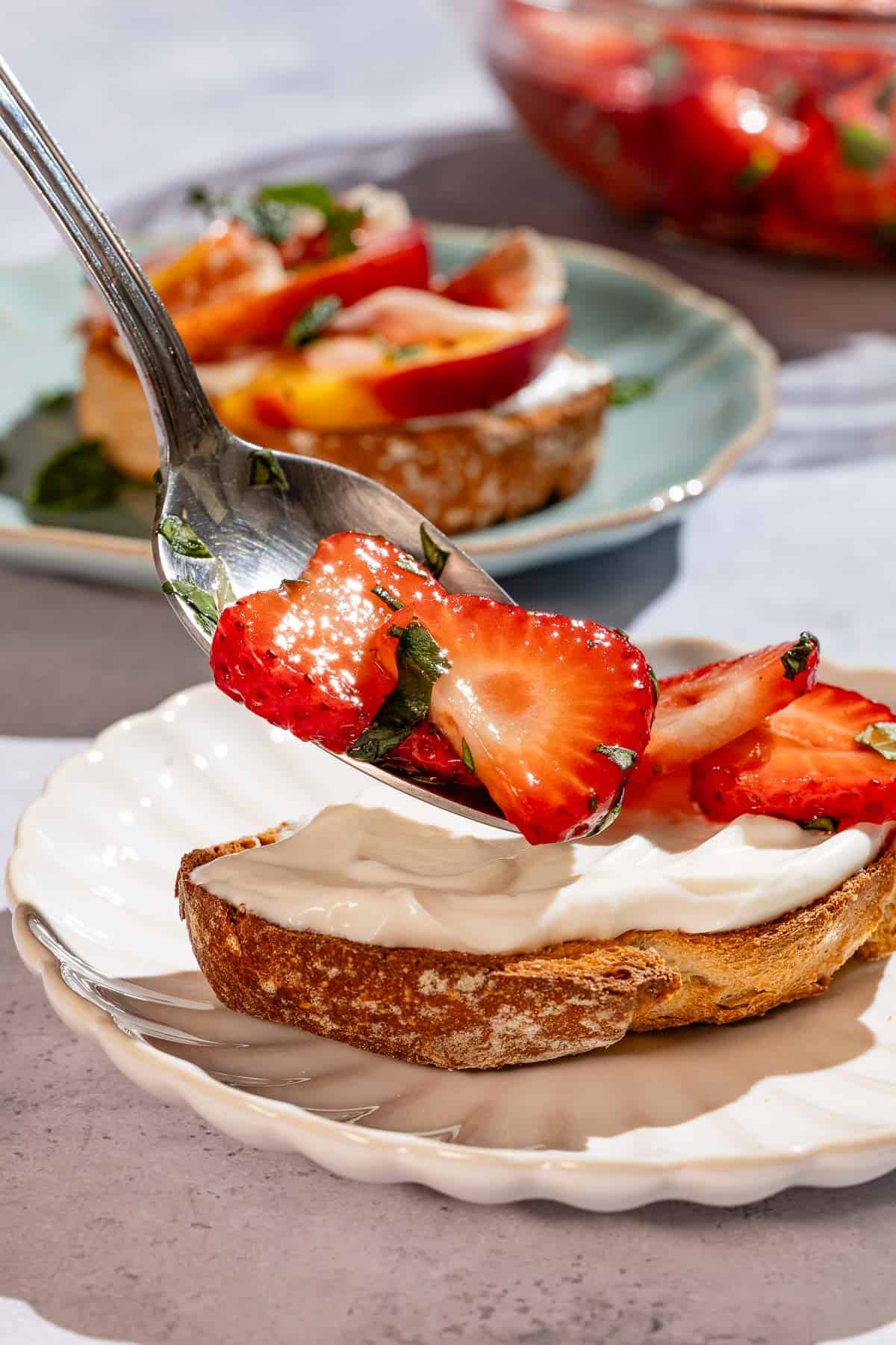 A close up of ricotta toast being topped with macerated strawberries. In the background is another ricotta toast.