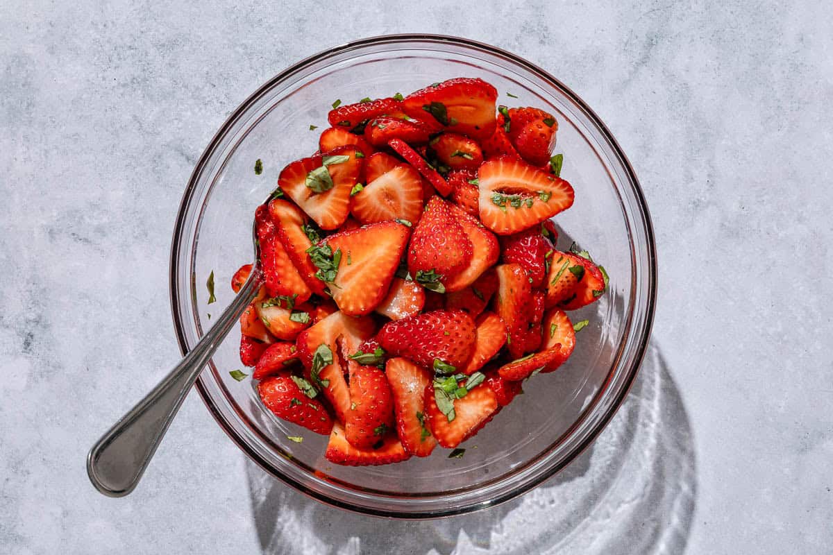 Sliced strawberries in a bowl with balsamic vinegar, honey and basil, as well as a spoon.