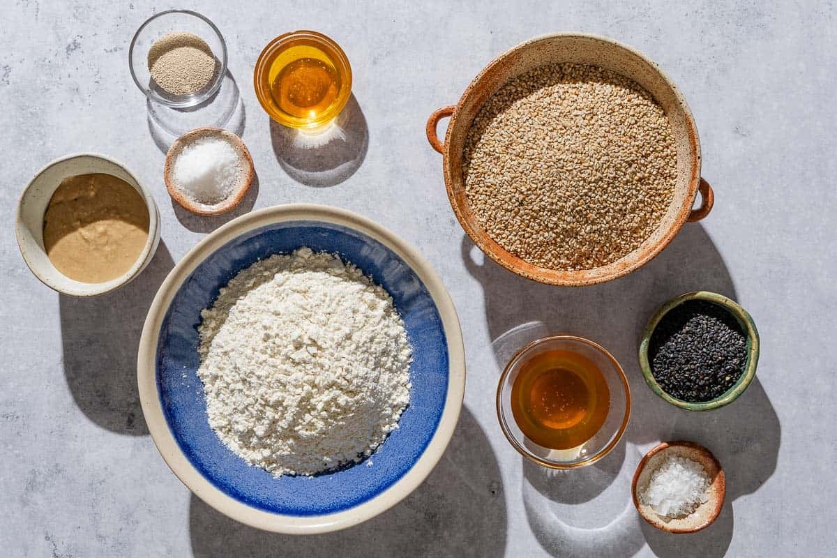 Ingredients for crispy sesame breadsticks including instant dry yeast, honey, tahini paste, all-purpose flour, kosher salt, white sesame seeds, black sesame seeds, and sea salt.