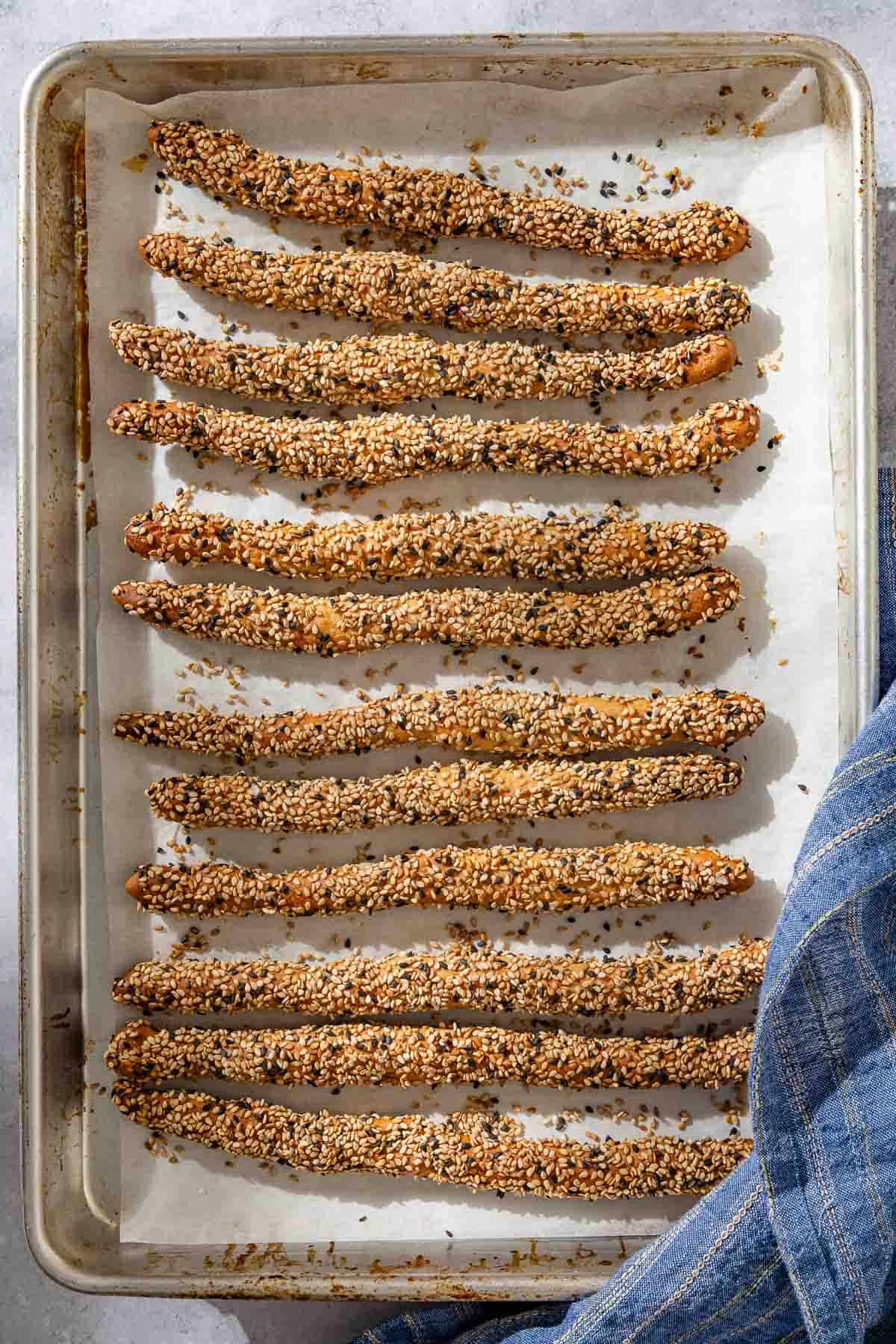 An overhead photo of 12 baked crispy sesame breadsticks on a parchment lined sheet pan next to a kitchen towel.