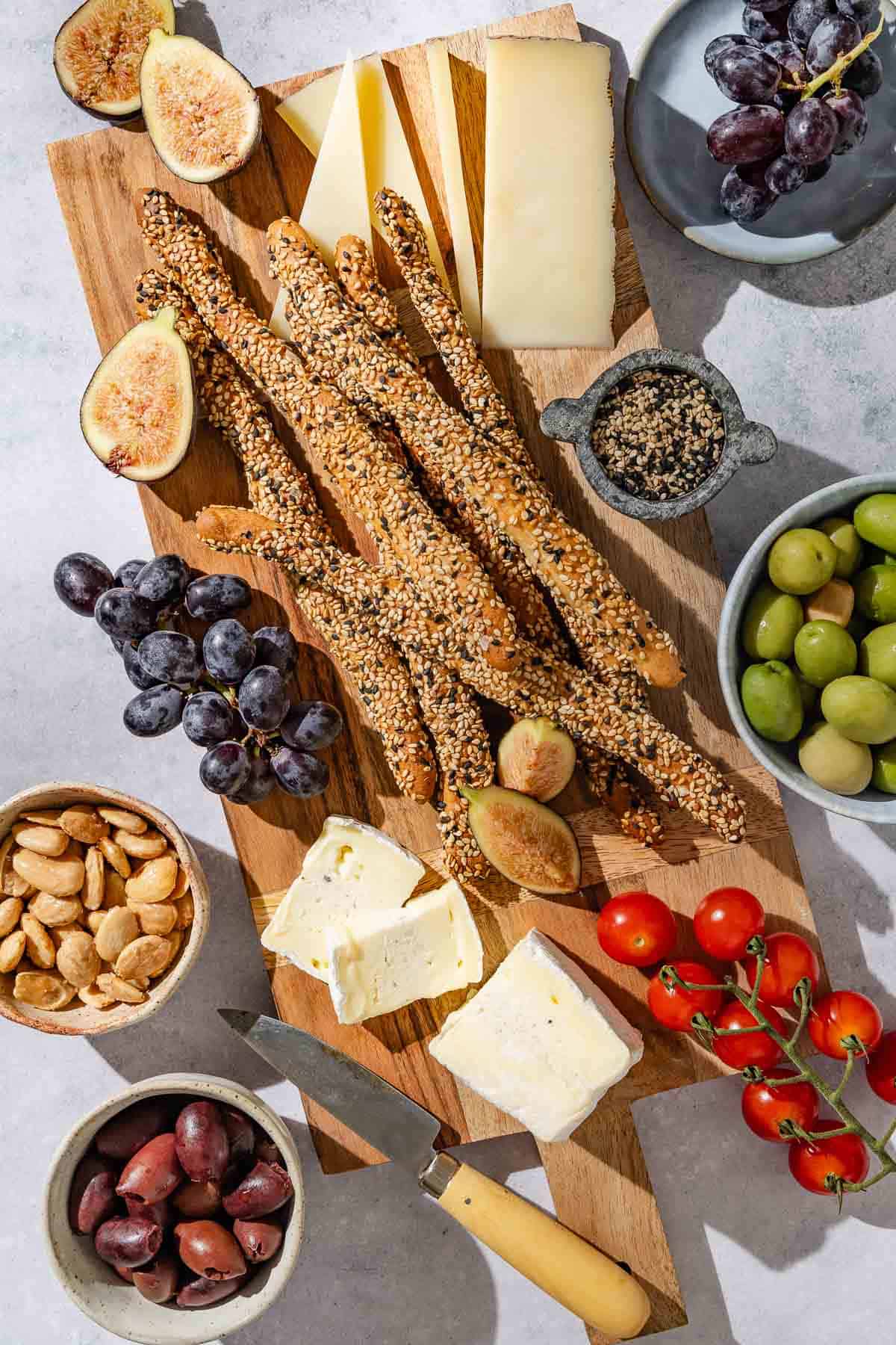 Several baked crispy sesame breadsticks on a wooden charcuterie board with various fruits, nuts, olives, cheeses and a knife.