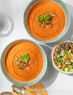 Overhead photo of 2 bowls of watermelon gazpacho topped with, diced watermelon, cucumber, green pepper, olive oil, black pepper and mint next to a bowl of these garnishes and two spoons.