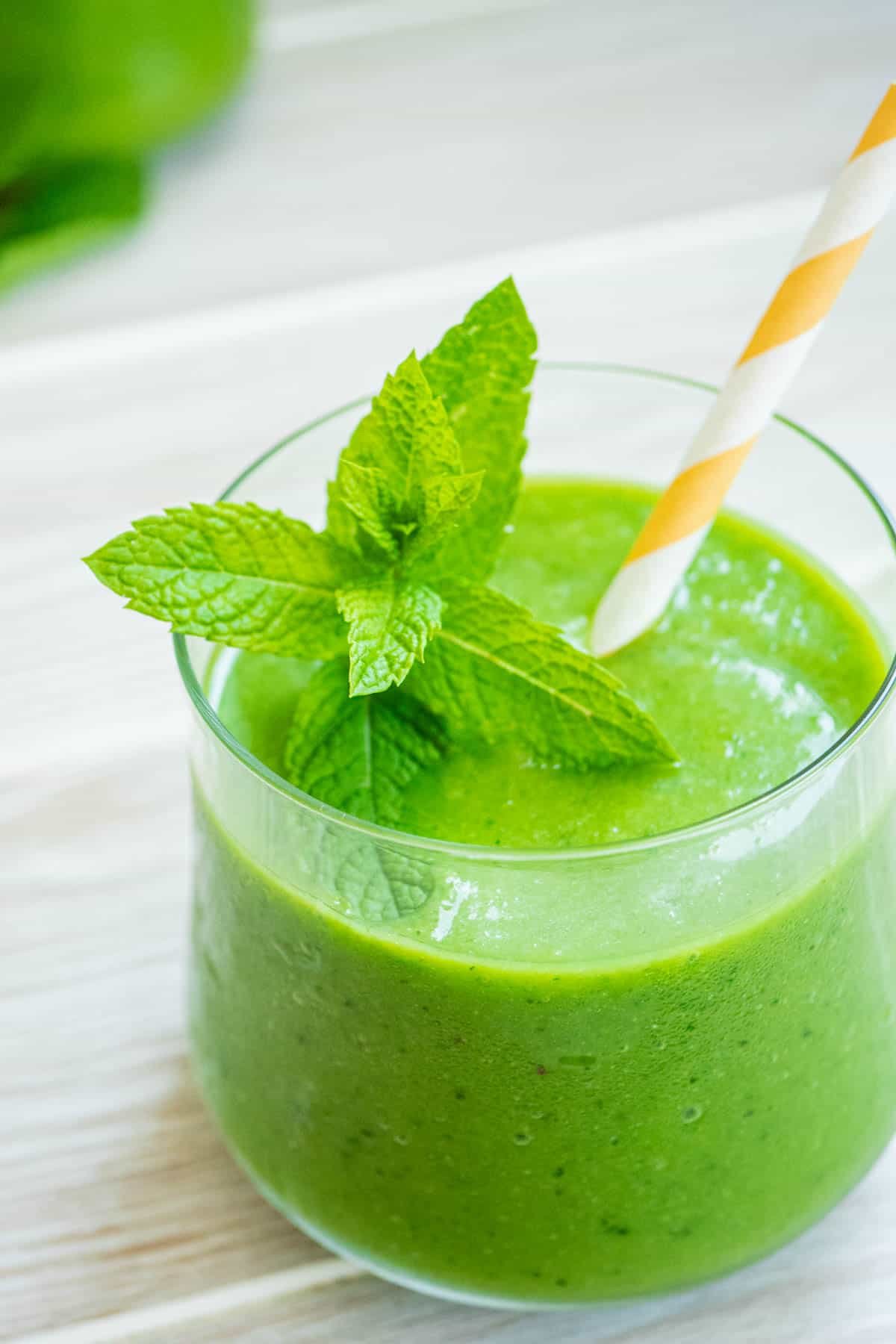 a vegan green smoothie in a glass with fresh mint and a yellow and white straw.