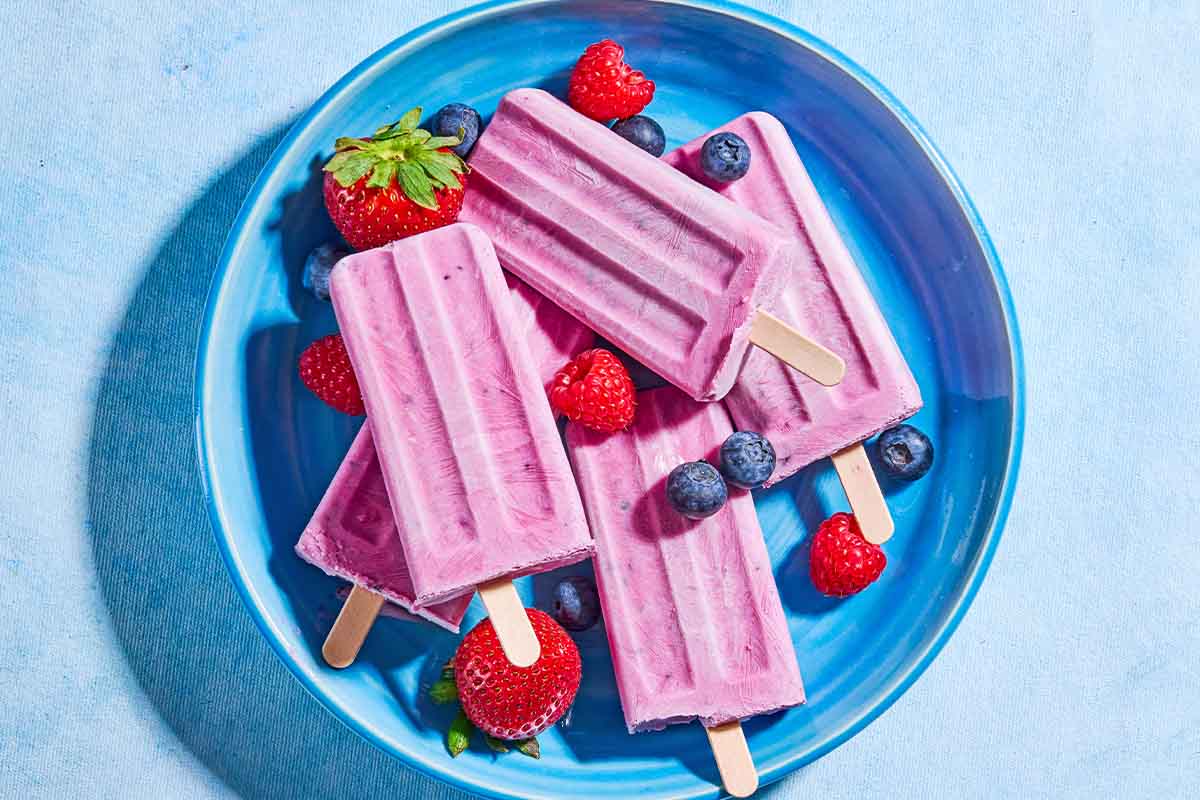 An overhead photo of 4 yogurt berry homemade popsicles surrounded by blueberries, strawberries and raspberries on a plate.