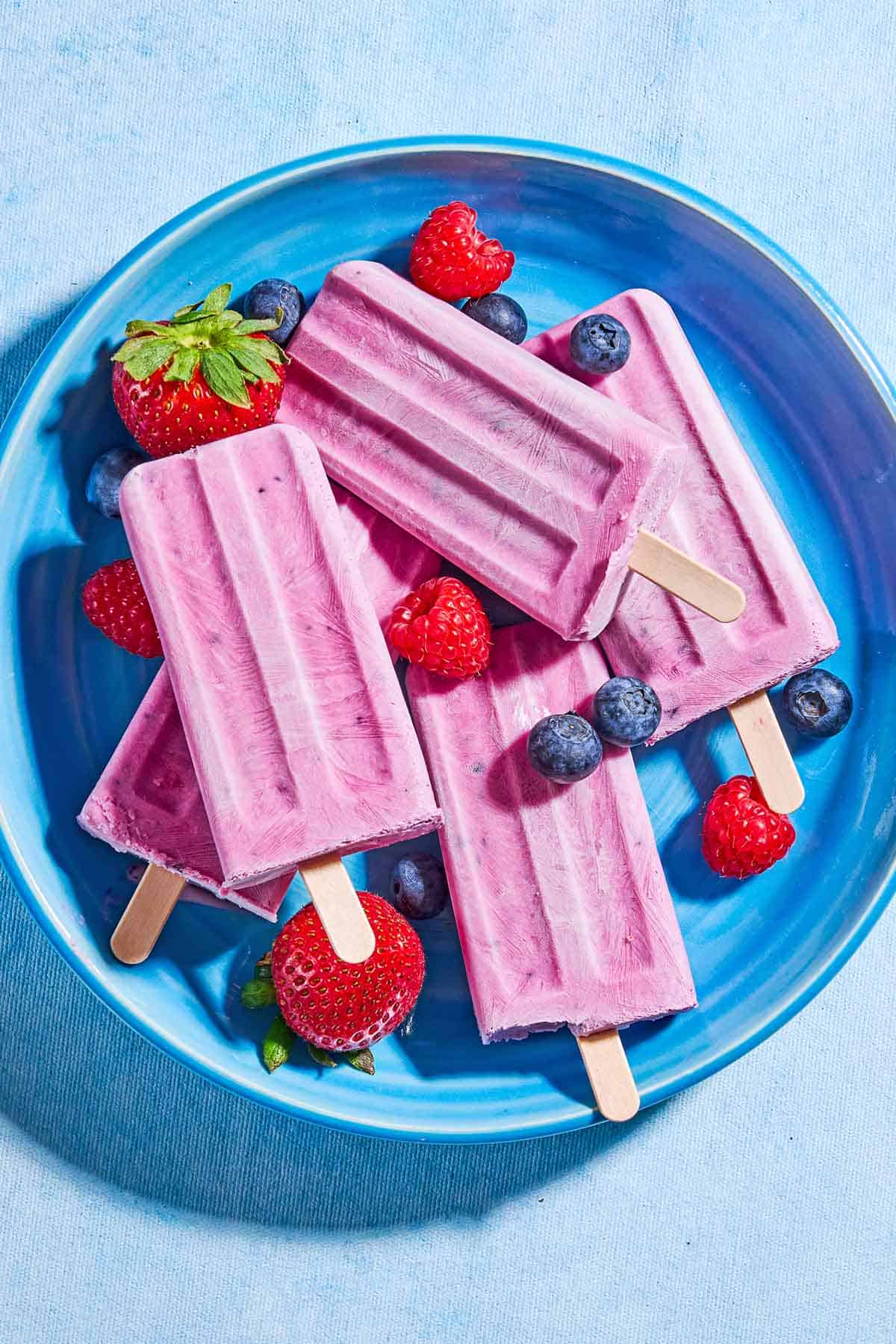 An overhead photo of 4 yogurt berry homemade popsicles surrounded by blueberries, strawberries and raspberries on a plate.