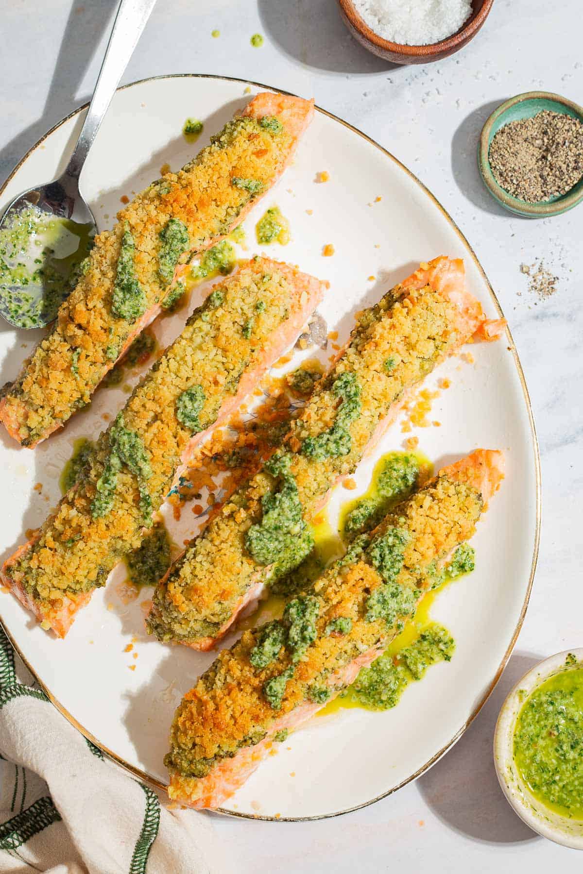 An overhead photo of 4 pesto salmon fillets on a platter with a spoon. Next to this are bowls of salt, pepper and pesto.