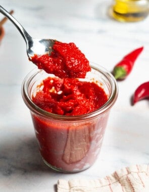 Red pepper paste being lifted out of a jar of the paste on a spoon. Surrounding this is a bowl of olive oil, two red chilies and a kitchen towel.