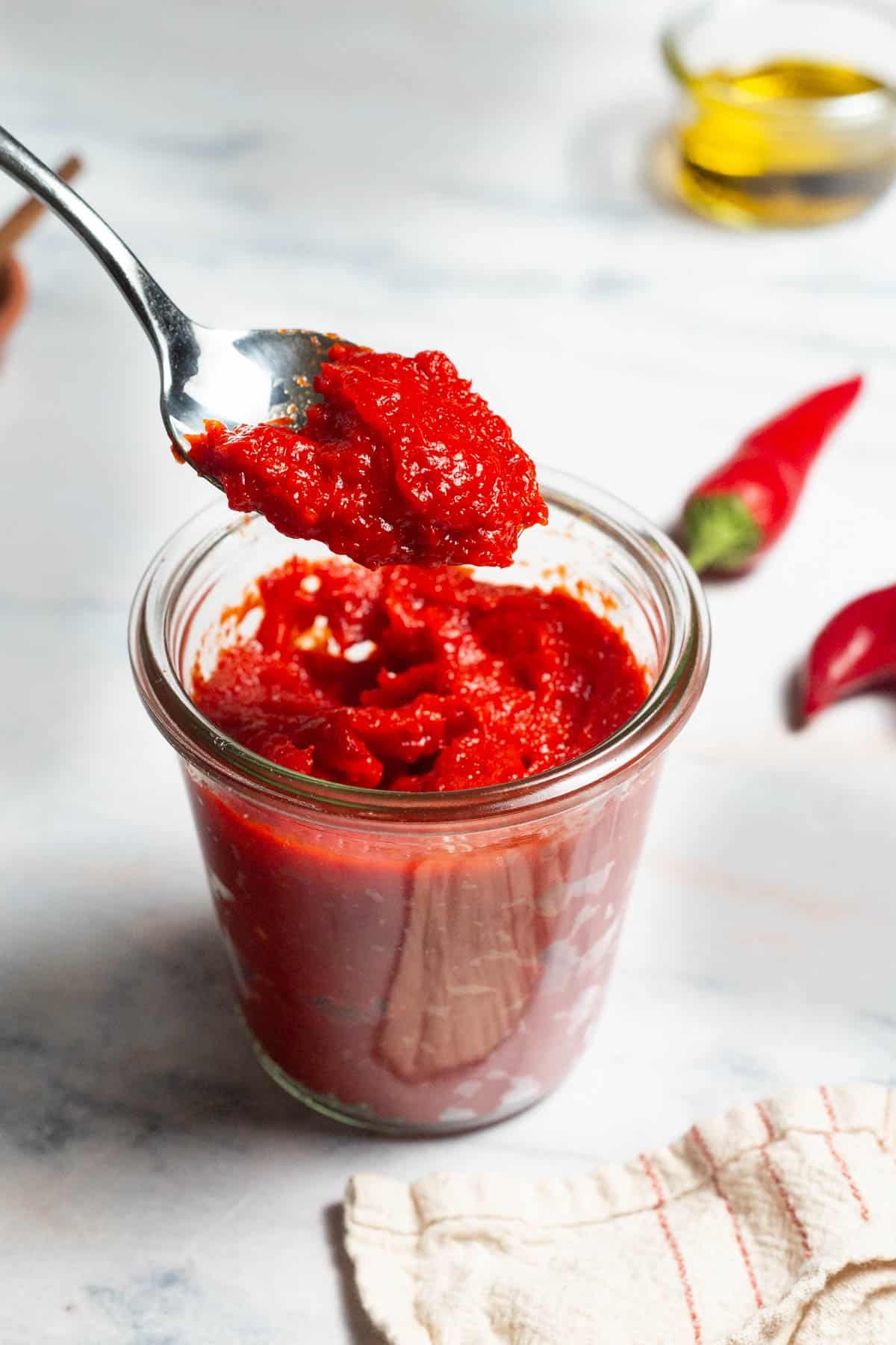 Red pepper paste being lifted out of a jar of the paste on a spoon. Surrounding this is a bowl of olive oil, two red chilies and a kitchen towel.