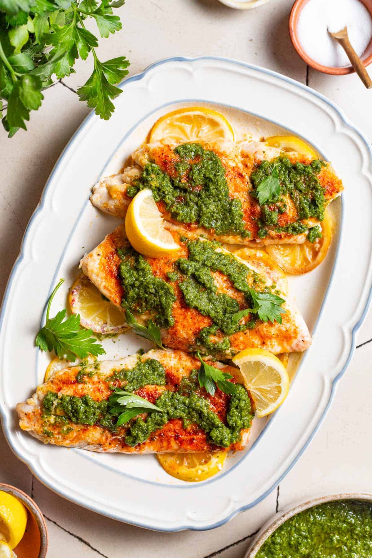 An overhead photo of 3 baked red snapper fillets topped with zhoug and parsley laying on lemon rounds on a serving platter. Surrounding this is a bunch of parsley, and bowls of salt, pepper, zhoug and lemon slices.