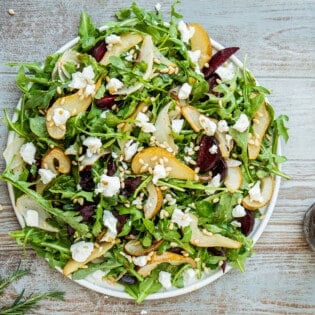 beet and goat cheese salad on a serving platter next to a jar of honey balsamic vinaigrette with a whisk and a bowl of pine nuts.