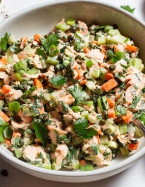 A close up of salmon salad in a serving bowl with a spoon next to bowls of pepper, parsley and capers, and a kitchen towel.