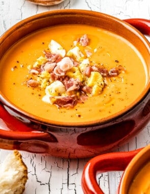 close up of a bowl of salmorejo spanish cold tomato soup topped with chopped hard boiled eggs and prosciutto, next to another bowl of soup, some crusty bread, a glass of wine, and a bowl chopped eggs and prosciutto.