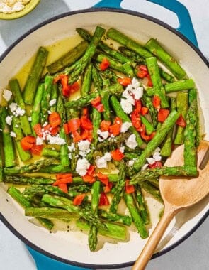 Sauteed asparagus topped with chopped red peppers and crumbled feta in a skillet with a wooden fork. Next to this is a bowl of crumbled feta.