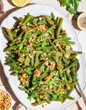 An overhead photo of sauteed green beans on a serving platter with a spoon. Surrounding this is a lemon half, kitchen towel, parsley, and bowls of salt, black pepper and pine nuts.