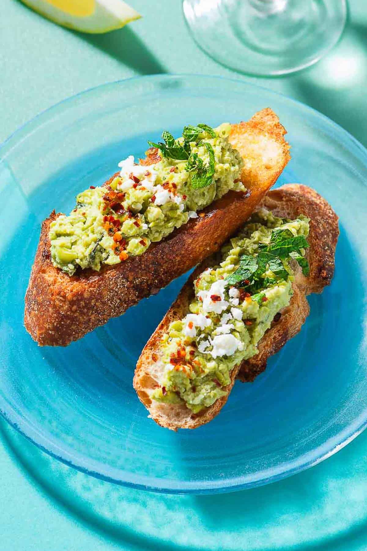 A close up photo of 2 pieces of smashed peas on toast garnished with aleppo pepper, feta and mint on a plate.