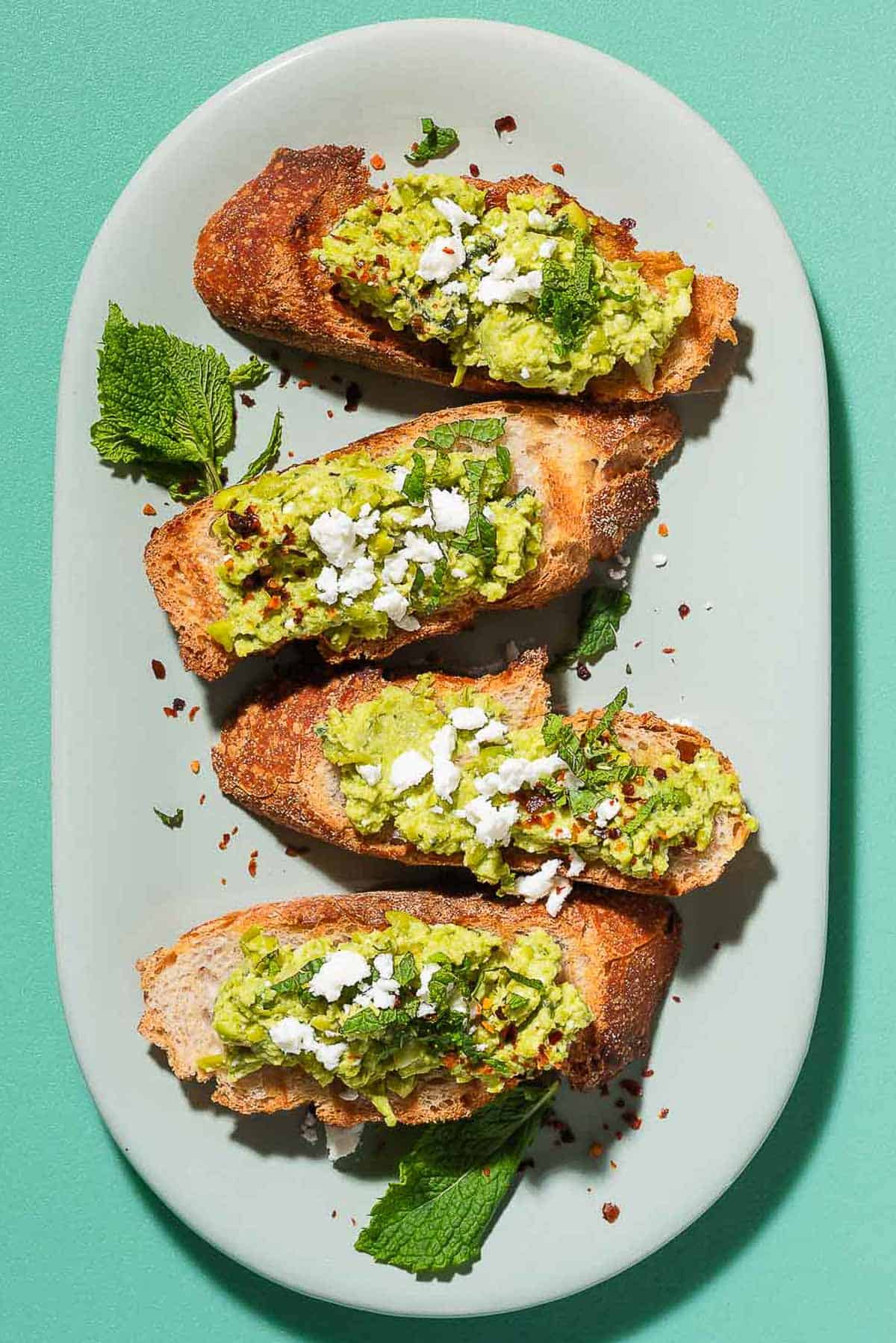 An overhead photo of 4 pieces of smashed peas on toast garnished with aleppo pepper, feta and mint on a serving platter.