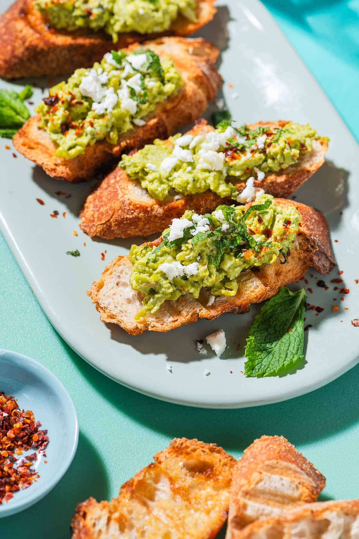 A close up photo of 4 pieces of smashed peas on toast garnished with aleppo pepper, feta and mint on a serving platter. Next to this is a small bowl of Aleppo pepper, and 3 pieces of tasted bread.